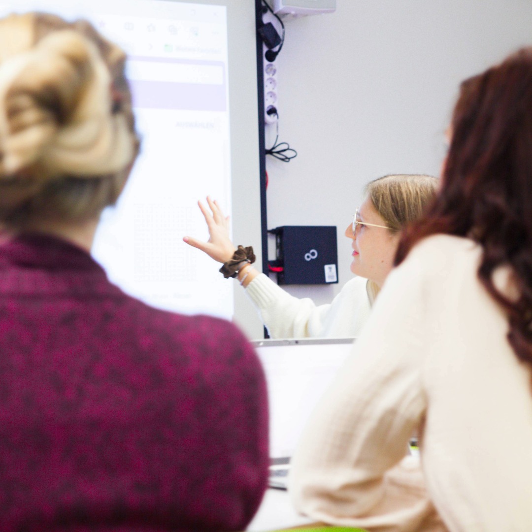 Zwei Personen von hinten sitzen vor einem Bildschirm, eine dritte Person zeigt mit der Hand auf den Bildschirm